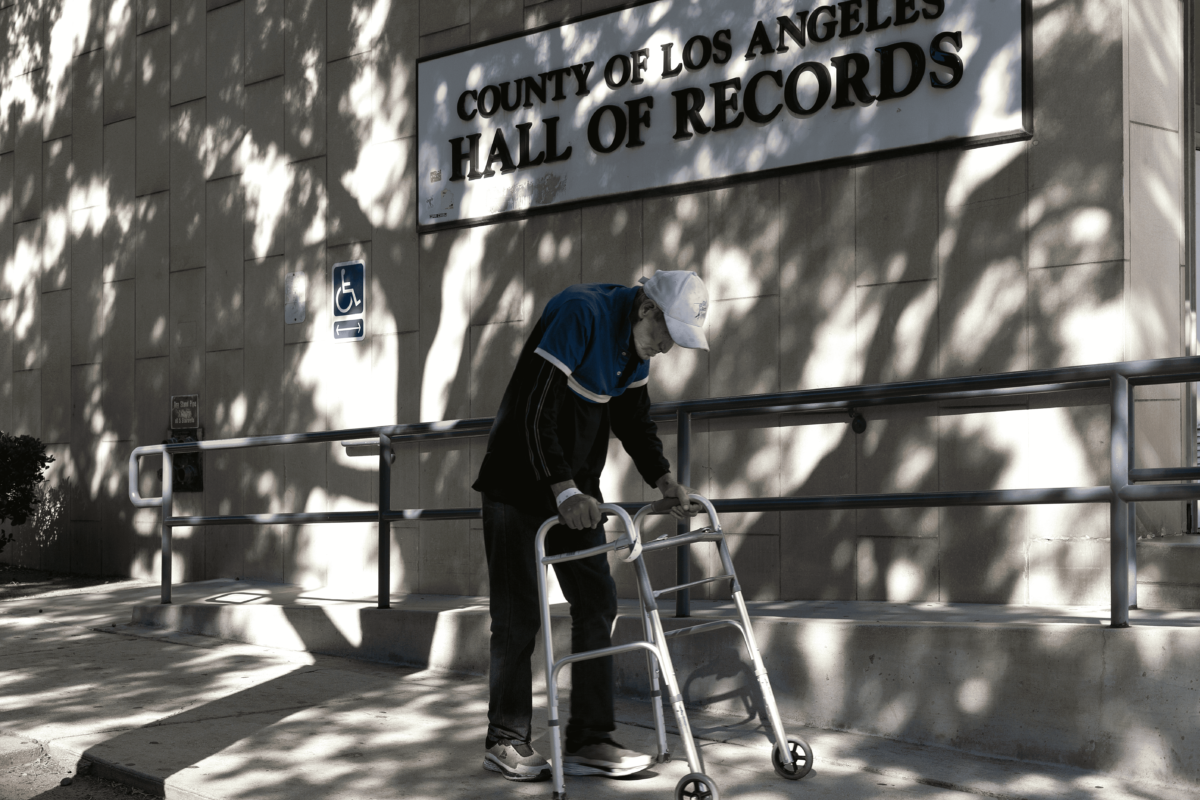 An elderly man and his walking device, shadowed by trees. On the wall behind a sign reads utside the L.A. County Hall of Records. 